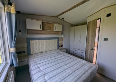 Master bedroom neutral wooden and yellow tones, master bed with overhead and bedside cabinets, wardrobe and drawer, with door leading into the en suite shower room