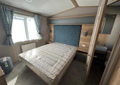 Master bed with a large rustic warm wooden panel behind the bed and padded navy headboard attached. Bedside cabinets and lighting, overhead storage, radiator and blue and grey floral curtains looking on to the en suite wc